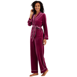 A woman models a two-piece pajama set with a long-sleeve, V-neck top and matching pants in a deep burgundy velvet material, accented with a contrasting trim and waist tie.