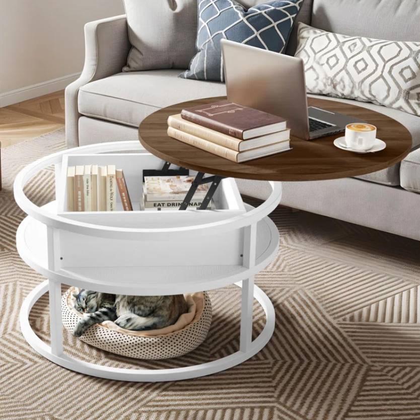 A round, white coffee table with a lift-top wooden surface holds books and a laptop. Below, a cat rests in a cushioned bed on a patterned rug.