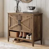 A rustic wooden shoe cabinet with black handles features a shelf holding boots, sneakers, and a wicker basket, topped with decor items like books, a bowl, and a glass vase with branches, set on a light wooden floor next to a textured rug.