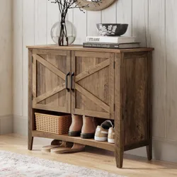 A rustic wooden shoe cabinet with black handles features a shelf holding boots, sneakers, and a wicker basket, topped with decor items like books, a bowl, and a glass vase with branches, set on a light wooden floor next to a textured rug.