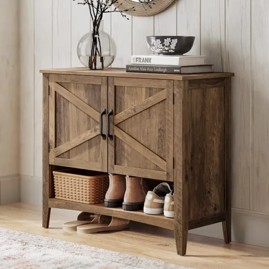 A rustic wooden shoe cabinet with black handles features a shelf holding boots, sneakers, and a wicker basket, topped with decor items like books, a bowl, and a glass vase with branches, set on a light wooden floor next to a textured rug.