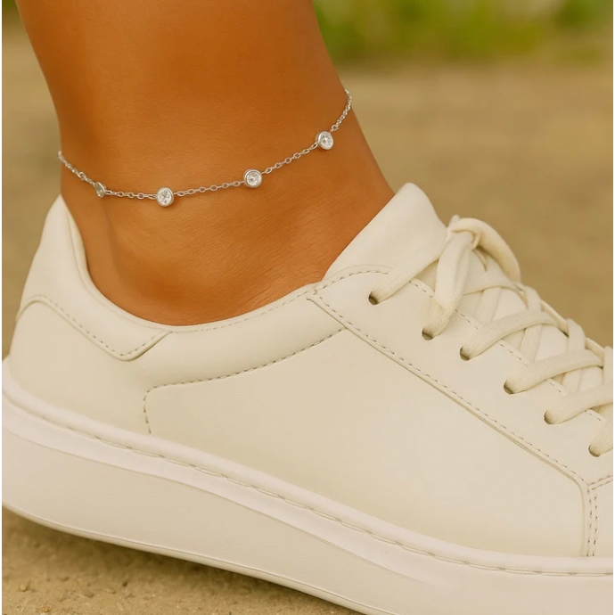 A delicate silver anklet featuring a chain with evenly spaced crystal embellishments. It is worn around the ankle and paired with a white sneaker.