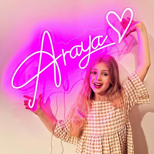 A young girl poses with a pink neon sign that spells \“Araya\“ with a heart. She is wearing a checkered dress and smiling.