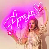 A young girl poses with a pink neon sign that spells \“Araya\“ with a heart. She is wearing a checkered dress and smiling.