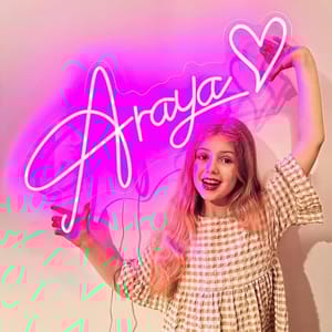 A young girl poses with a pink neon sign that spells \“Araya\“ with a heart. She is wearing a checkered dress and smiling.