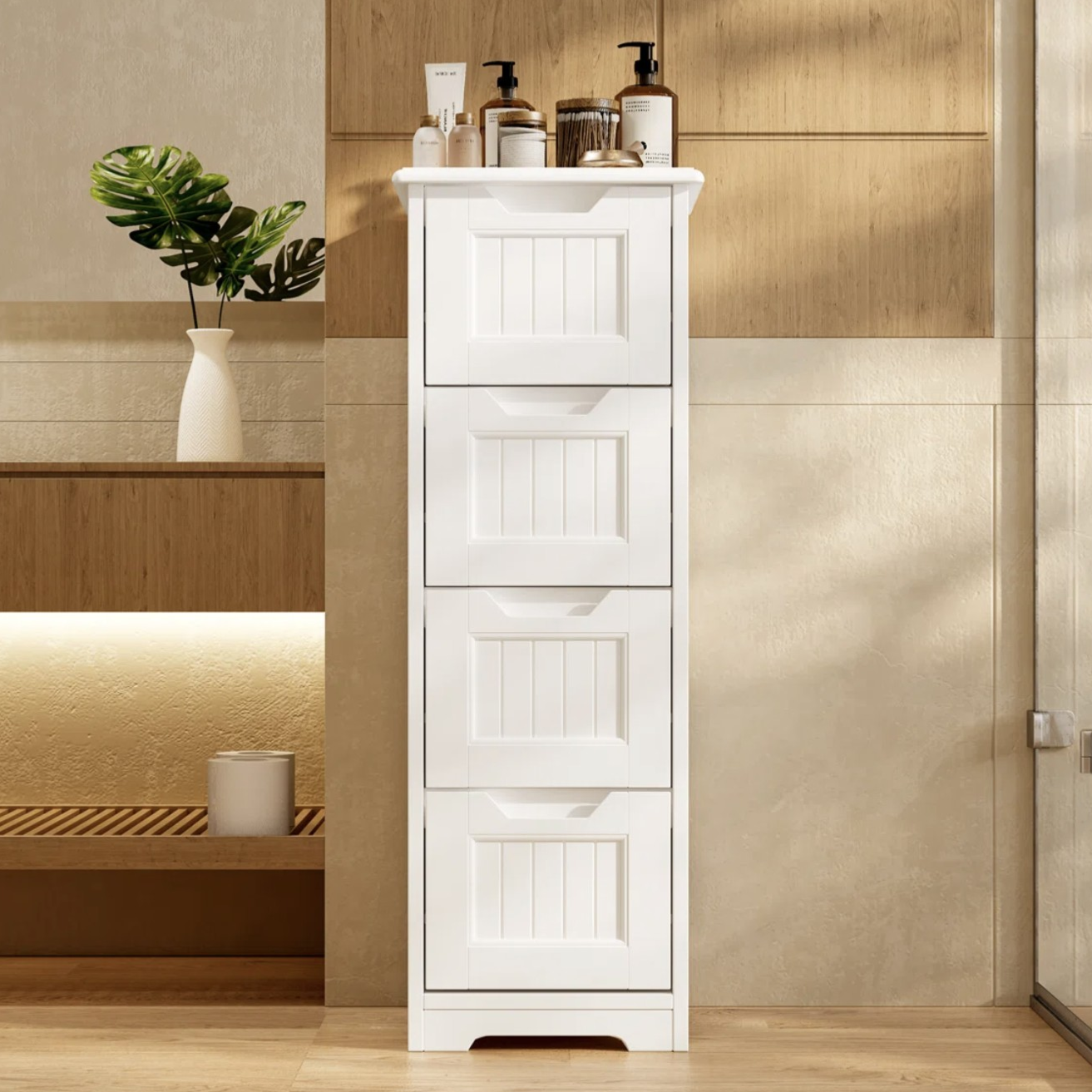 A white, tall storage cabinet with multiple drawers stands against a wooden wall in a room. The top is adorned with various toiletry items, and there's a decorative vase with green leaves nearby.