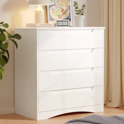 A white four-drawer dresser is topped with a lamp, digital clock, and framed artwork. A potted plant is nearby, and the setup is against a light-colored curtain.