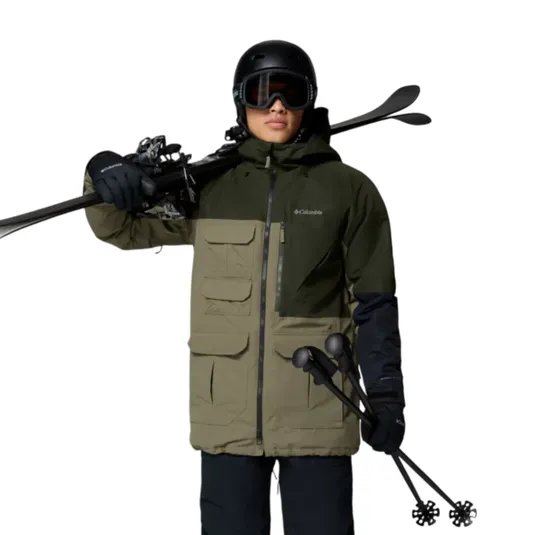 A person dressed in winter sports gear holds skis and poles, wearing a Columbia jacket, snow pants, gloves, boots, and a helmet with goggles. The outfit is designed for skiing, featuring a green and black jacket with multiple pockets.
