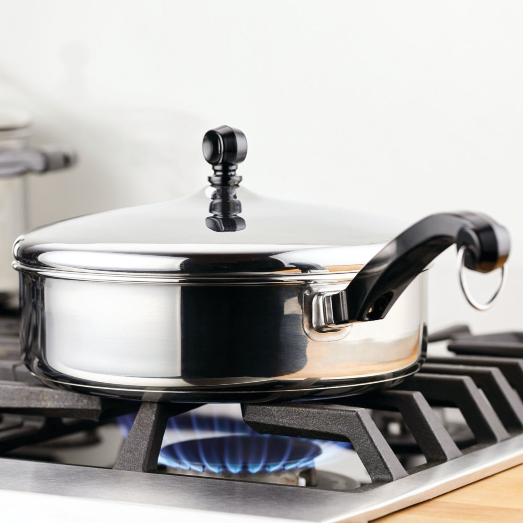 A stainless steel saucepan with a lid and ergonomic handle rests on a gas stove, with a visible blue flame beneath.