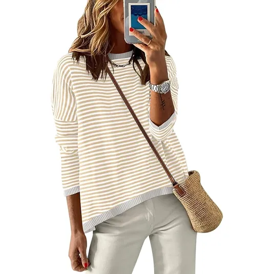A woman is wearing a cream and white striped long-sleeve top, white pants, a watch, and a necklace. She carries a straw crossbody bag and holds a smartphone with red nail polish visible.