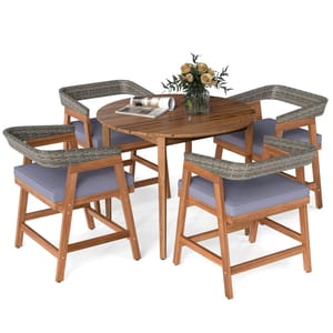 Round wooden dining table with four chairs featuring woven backrests and gray cushions, adorned with a vase of flowers and magazines.