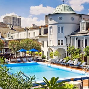 A luxurious outdoor pool area with lounge chairs and blue umbrellas, set within a scenic urban hotel complex featuring charming, traditional-style architecture.