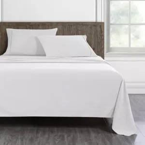 A bed with a wooden headboard is made with white sheets, two white pillows, and a smooth white blanket, situated in a room with a large window and wooden flooring.