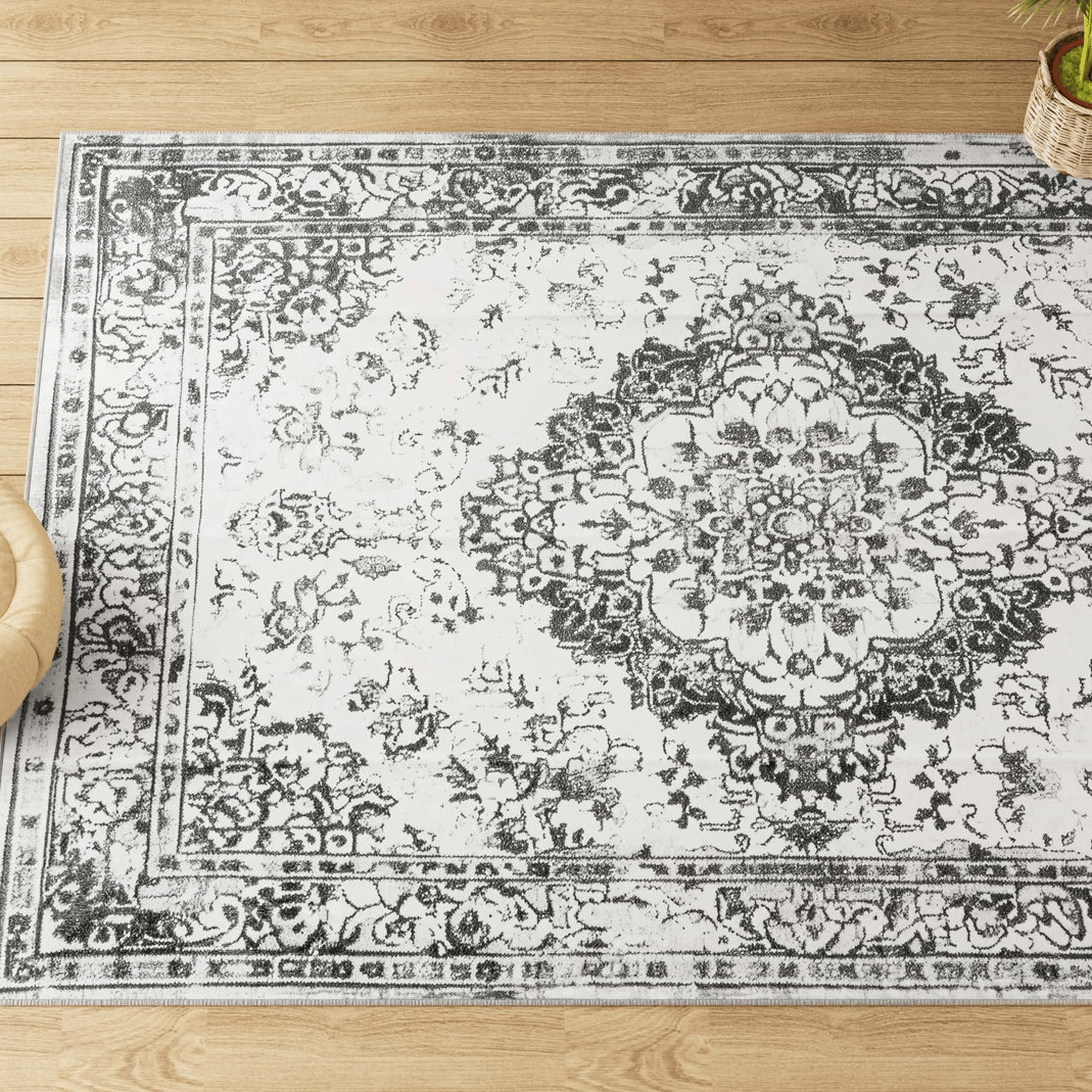 A vintage-style area rug with a distressed black and white floral medallion pattern on a pale background is placed on a wooden floor, accompanied by a small plant.