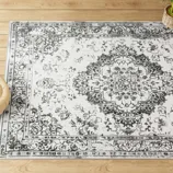 A vintage-style area rug with a distressed black and white floral medallion pattern on a pale background is placed on a wooden floor, accompanied by a small plant.