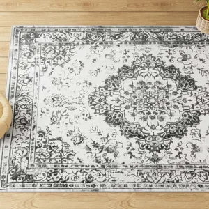 A vintage-style area rug with a distressed black and white floral medallion pattern on a pale background is placed on a wooden floor, accompanied by a small plant.