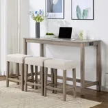 A wooden console table with a power outlet on the side is accompanied by three padded, beige stools. The table is decorated with a vase of blue flowers, a laptop, and a mouse, set against a white wall with framed floral artwork.