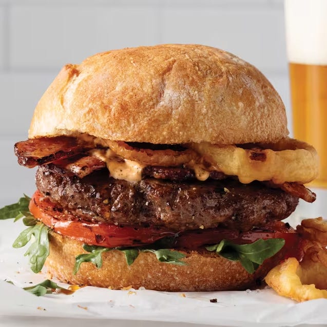 A burger with a beef patty, bacon, arugula, tomato, and onion rings on a round bun, with a creamy sauce visible. A glass of beer is in the background.