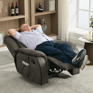 A man relaxes on a gray recliner chair with a control panel, side pocket, and cushioned features, positioned in a living room with a bookshelf holding wine bottles and decor.