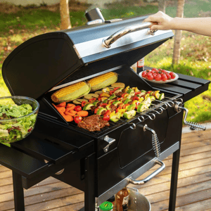 A grill is loaded with various foods, including corn, skewers with vegetables and meat, and a steak. A bowl of salad and a plate of tomatoes are nearby, set on a patio deck in an outdoor setting.