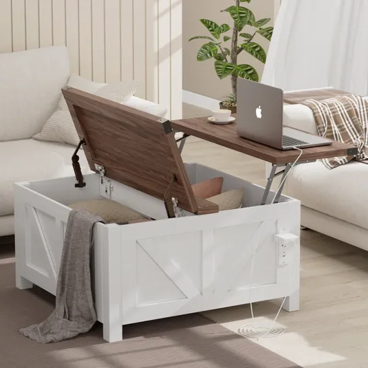 White wooden storage trunk with a lift-top table, revealing interior storage space, holding pillows and a blanket. A laptop and cup rest on the extended tabletop. A power outlet is mounted on the side.