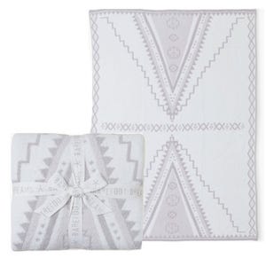 A soft Barefoot Dreams throw blanket with a geometric pattern in light gray and white, wrapped with a branded ribbon. The design features large V-shaped motifs with a southwestern style.