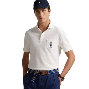 A man is wearing a white polo shirt with a polo bear patch, blue pants with a striped belt, and a navy cap featuring a small logo.