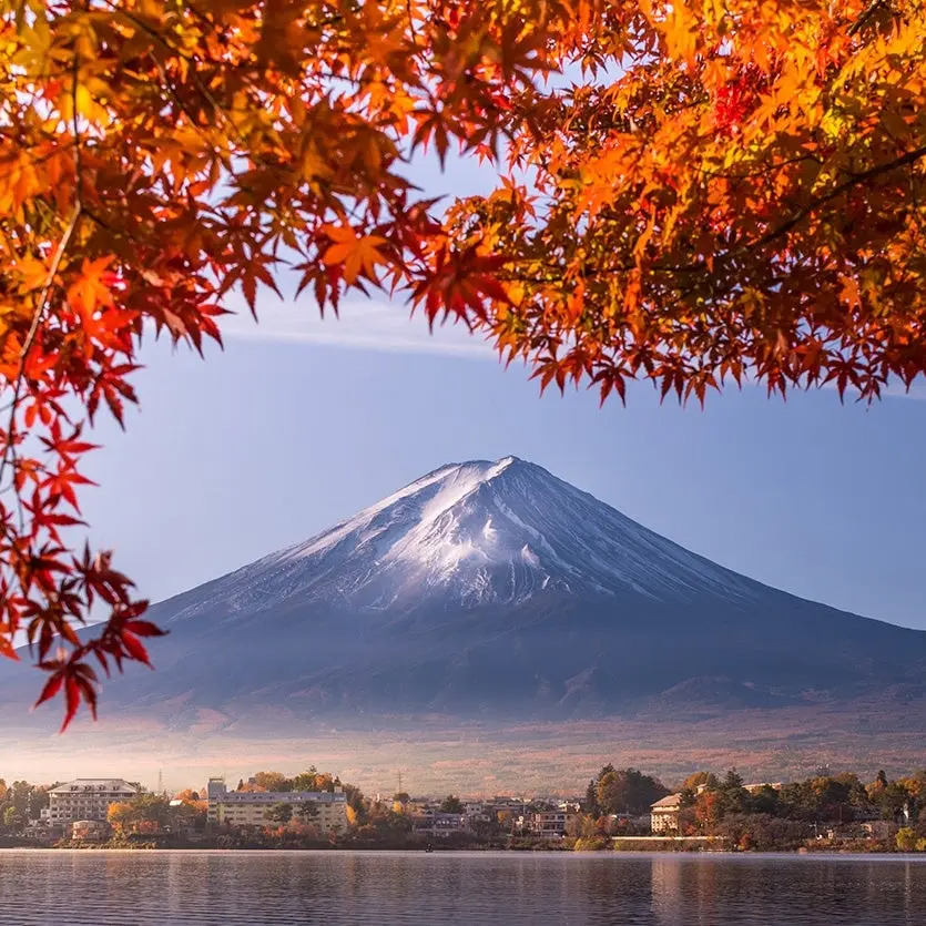 Snow-capped mountain with vibrant autumn foliage framing the scene, overlooking a calm lake and town buildings below.