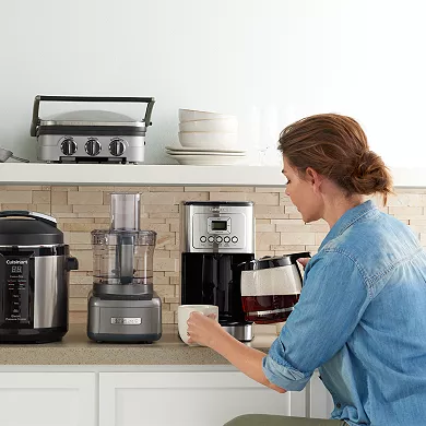 A woman is pouring a cup of coffee from a Cuisinart 14-Cup Coffee Maker placed on a kitchen counter, alongside other kitchen appliances.