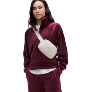 A woman wears a burgundy velour tracksuit with a high-neck top and matching pants, paired with a light-colored crossbody fleece bag.