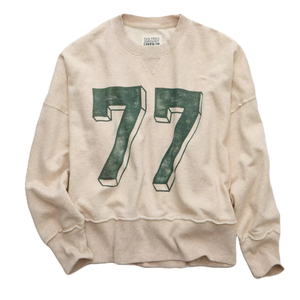 A beige long-sleeve sweatshirt featuring a large, stylized green \“77\“ on the front. The collar has a ribbed design and the label reads \“This Feels Seriously Lived In.\“