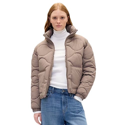 A woman wearing a puffy, taupe-colored jacket over a white turtleneck and blue jeans.