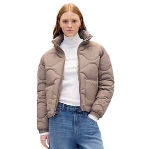 A woman wearing a puffy, taupe-colored jacket over a white turtleneck and blue jeans.