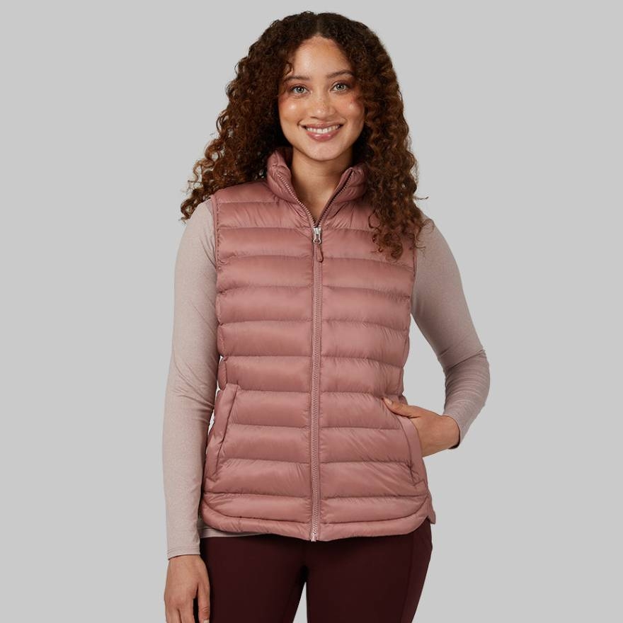 The vest is a sleeveless, zip-up design with a quilted pattern, in a muted pink shade, and has a high collar. The woman is pairing it with a long-sleeved top and maroon pants.