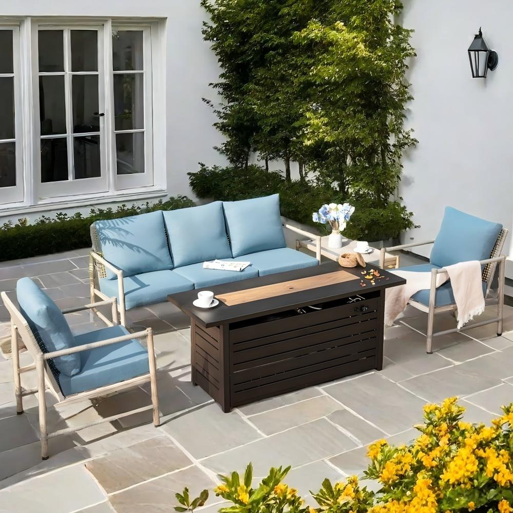 Outdoor patio set with light blue cushioned seating, including a sofa and two chairs, arranged around a rectangular, dark wood table. The setting is on a stone-paved area, with greenery and a building facade with windows in the background.