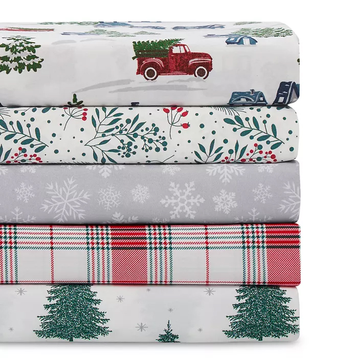 A stack of three twin-size holiday-themed sheet sets featuring patterns of a vintage truck, holly berries, snowflakes, plaid, and pine trees.