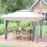 A beige outdoor gazebo with a mesh curtain encloses a wooden dining table and chairs, set on a wooden deck. A traditional brick house and a garden with flowers and greenery are visible in the background.