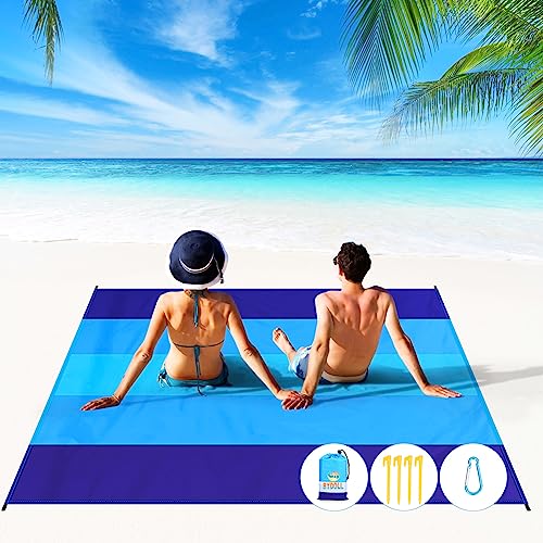 An oversized blue beach blanket is laid out on the sand with two people sitting on it, surrounded by a tropical beach scenery; stakes are included to secure it.