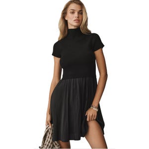 A woman is wearing a black dress with a ribbed, short-sleeve turtleneck top and a flowing skirt. She holds a patterned handbag and wears gold bracelets.