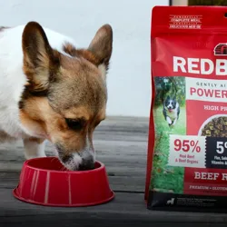 A dog eats from a red bowl next to a bag of Redbarn Powerfood, a high protein, air-dried dog food made with 95% real beef and beef organs, and 5% flax, salmon oil, vitamins, and minerals.