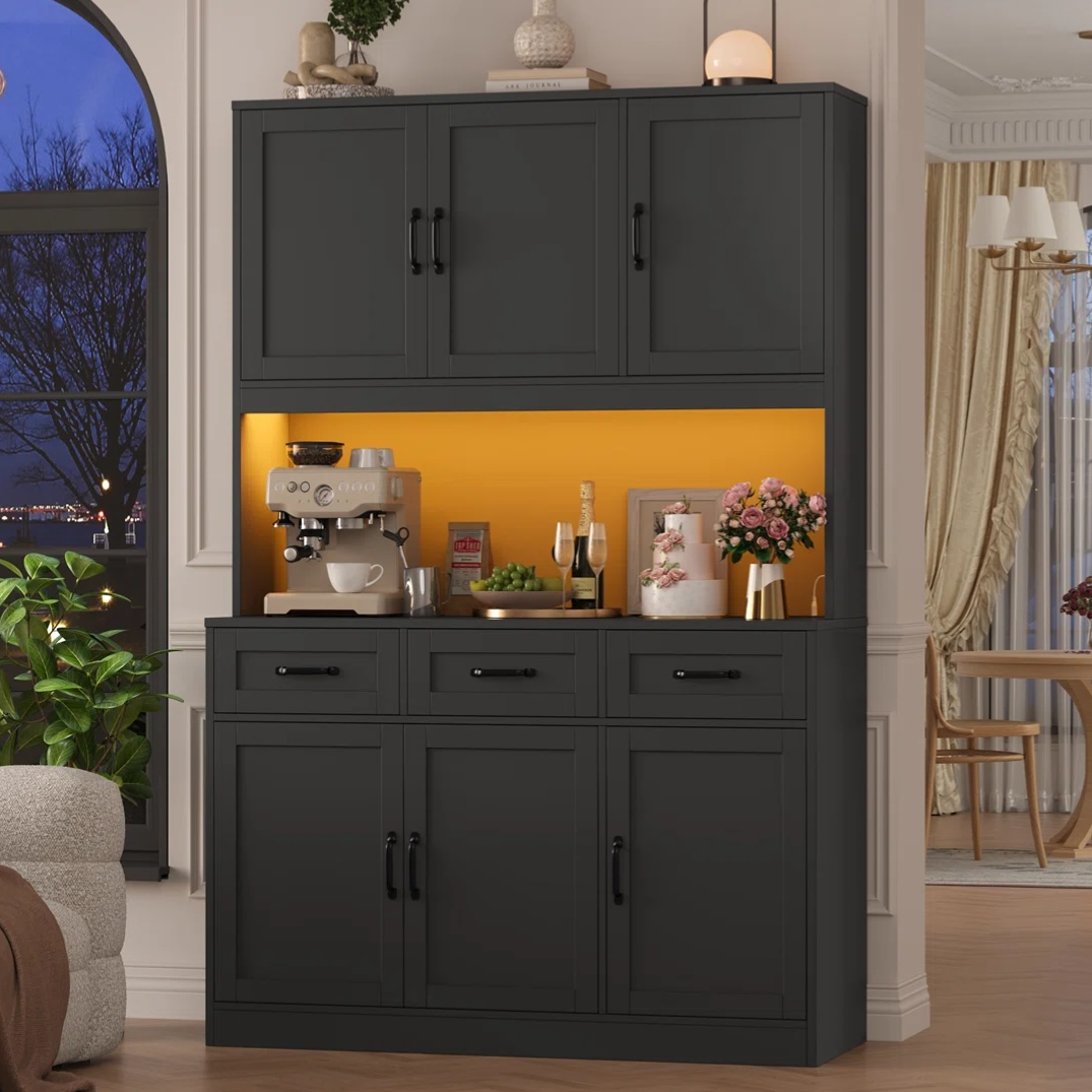 A black cabinet with upper and lower storage features a brightly lit yellow section containing a coffee maker, a cup, a wine bottle, a plant, and decorative items.