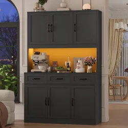 A black cabinet with upper and lower storage features a brightly lit yellow section containing a coffee maker, a cup, a wine bottle, a plant, and decorative items.