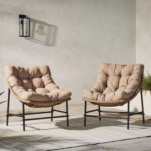 Two Papasan-style outdoor patio scoop chairs with woven rattan backs and plush, tufted beige cushions are shown on black metal frames, set on a striped outdoor rug against a textured wall.