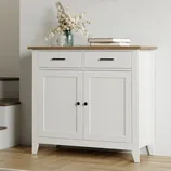 A white sideboard with a wooden top, two drawers, and two doors, adorned with a vase of dried flowers and books.