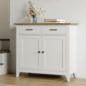 A white sideboard with a wooden top, two drawers, and two doors, adorned with a vase of dried flowers and books.