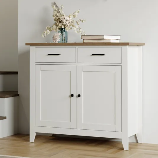A white sideboard with a wooden top, two drawers, and two doors, adorned with a vase of dried flowers and books.