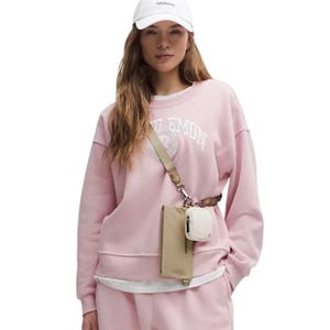 A woman wears a pink sweatshirt and matching pants with a beige lululemon Dual-Pouch Sling crossbody bag. The bag has two compartments, one with a zipper and another smaller pouch attached, and she's also wearing a white cap.