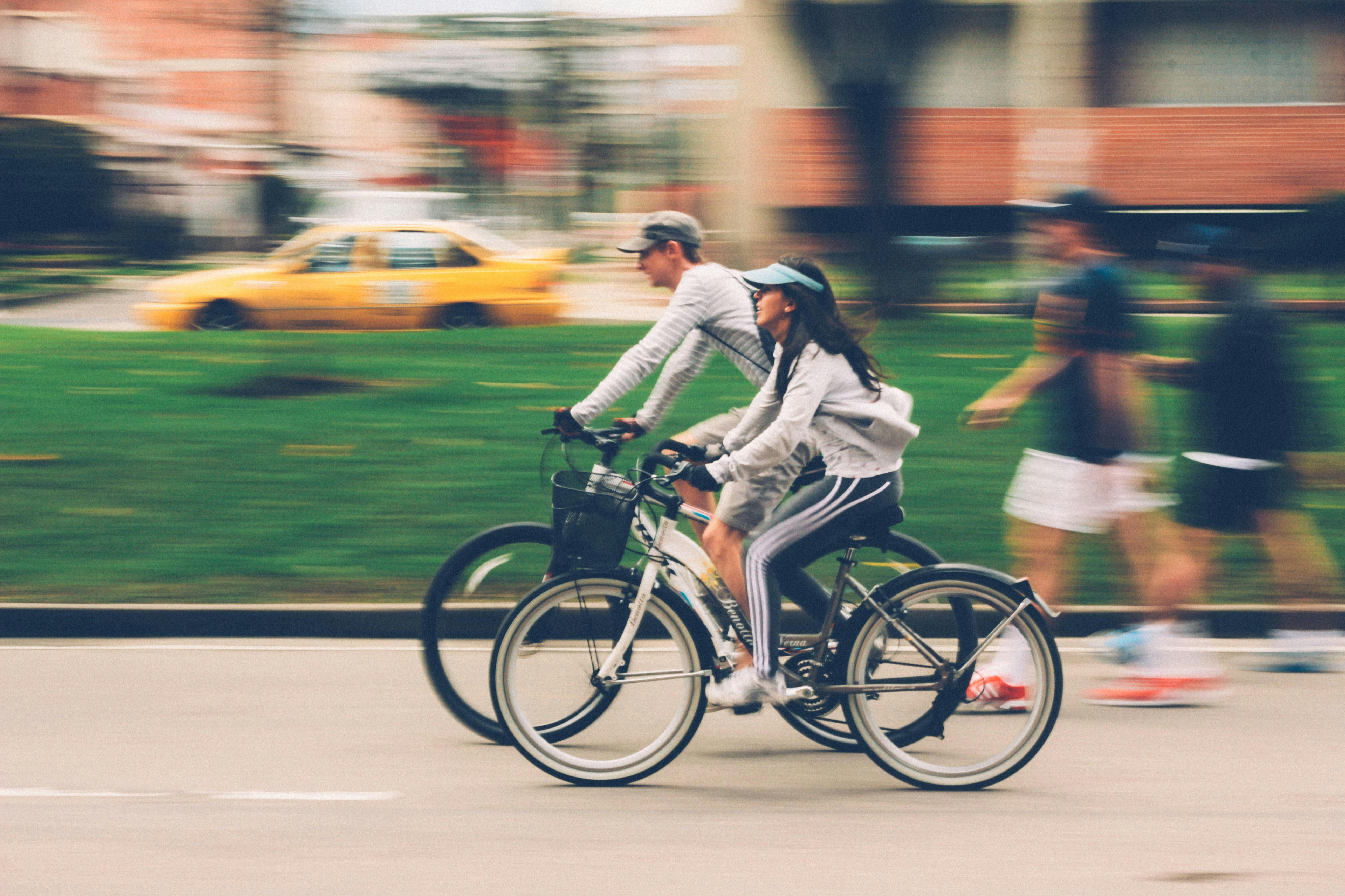 Two people riding bicycles, with motion blur suggesting movement, and a yellow taxi in the background.