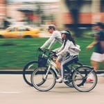Two people riding bicycles, with motion blur suggesting movement, and a yellow taxi in the background.