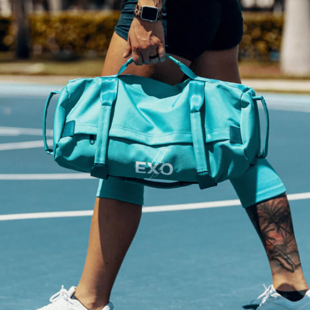 A person carries an aqua-blue EXO sports duffel bag while walking on a basketball court, wearing a smartwatch and shorts, with a tattoo on their calf.